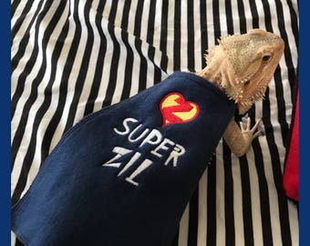 May include: A bearded dragon wearing a navy blue cape with the words "SUPER ZIL" embroidered in white. The cape features a red heart with a yellow lightning bolt. The background is a black and white striped pattern.