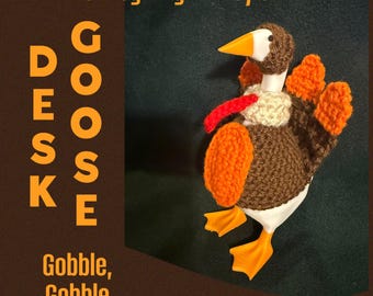 May include: A white plastic goose figurine wearing a brown, orange, and cream crocheted turkey costume. The text on the image reads: Thanksgiving Turkey outfit, DESK GOOSE, Gobble, Gobble. The costume includes a brown head cover, orange wings, and a red wattle.