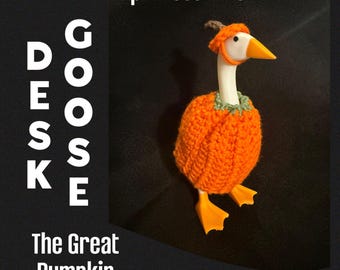 May include: A white plastic goose figurine wearing a crocheted orange pumpkin costume with a green collar and a small brown stem hat. The text "Pumpkin Costume", "DESK GOOSE", and "The Great Pumpkin" are printed on the black background.