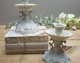 May include: Two white ceramic candle holders with gold trim and intricate designs. The holders are on a stack of books.