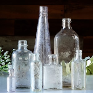 May include: A collection of antique glass bottles of various shapes and sizes. The bottles are clear with a frosted appearance, some with textured surfaces. The arrangement includes a tall, slender bottle and several shorter, rounded ones.