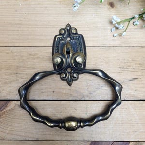 Puede incluir: Anillo para toalla de metal negro y dorado adornado con un diseño de volutas decorativas. El anillo está unido a un soporte de pared con una cerradura.