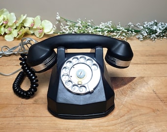Könnte beinhalten: Schwarzes Vintage-Wählscheibentelefon mit silbernem Zifferblatt und Akzenten. Das Telefon hat einen gebogenen Hörer und ein Spiralkabel. Das Telefon steht auf einer Holzoberfläche mit Blumen im Hintergrund.
