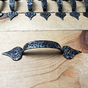 May include: Close-up of several hammered metal door pulls. The pulls are dark grey with a textured surface and a decorative, pointed end. The handles are arched and have a hammered texture. The background is a light-coloured wooden surface.