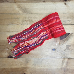 May include: A red woven scarf with a fringed edge. The scarf has a plaid pattern in red, white, and orange. The fringe is made of multiple colours, including red, white, yellow, green, and blue. The scarf is lying on a wooden surface.