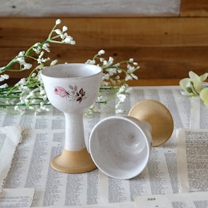 May include: Two handmade ceramic goblets. One upright, the other lying on its side. The goblets are speckled white with a tan base. One goblet has a pink rose and brown leaf design. The goblets are on a vintage paper background.