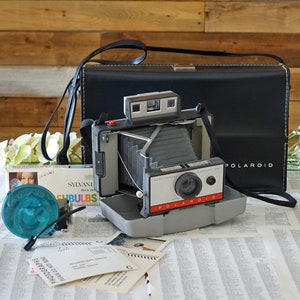 Peut inclure: Un appareil photo Polaroid Land vintage avec un étui en cuir noir et un flash. L'appareil photo est en bon état et présente un design argenté et noir. L'appareil photo est posé sur une table avec un journal et une boîte d'ampoules flash.