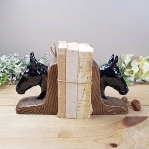 Op de afbeelding: Een paar decoratieve boekensteunen met zwarte paardenkoppen. De boekensteunen zijn gemaakt van keramiek en hebben een bruine, getextureerde basis. Een stapel boeken wordt tussen de boekensteunen gehouden, vastgebonden met touw.