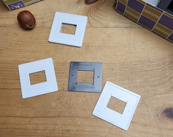 Può includere: Una collezione di supporti per diapositive Kindermann vintage e una singola nocciola su una superficie di legno. I supporti per diapositive sono in plastica bianca e un supporto in metallo grigio con il logo Kindermann. Scatole di supporti per diapositive sullo sfondo.