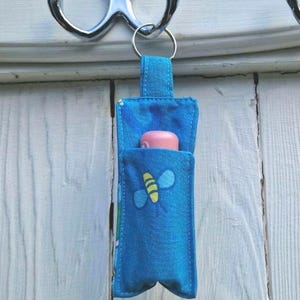 May include: A blue fabric keychain holder with a bee design, holding a pink lip balm. The holder is attached to a silver key ring and hangs from a silver hook. The bee has yellow and blue details.