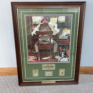 May include: Framed print featuring a man at a desk, head resting on arms, surrounded by papers. The print is in a dark brown frame with a green mat. Below the print are three postage stamps. A gold-colored nameplate is at the bottom.