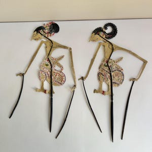 May include: Two traditional Indonesian shadow puppets, or wayang kulit, are displayed against a white background. The puppets feature intricate designs with tan, red, and black details. Each puppet has long, slender arms and legs, and is held by a black rod.