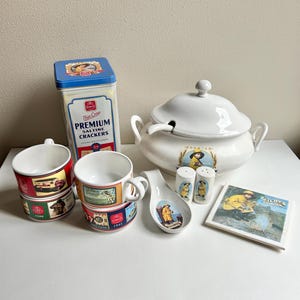 May include: A collection of vintage kitchenware featuring a white ceramic soup tureen with a lid, two mugs, a spoon rest, salt and pepper shakers, a coaster, and a tin of crackers. The items have a nautical theme with illustrations of a fisherman.