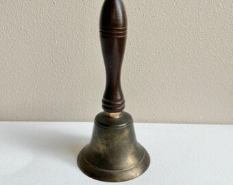 May include: A brass bell with a wooden handle. The bell has a gold-colored top and a dark brown handle.