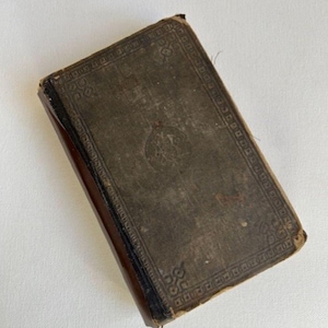 May include: A small, brown, leather-bound book with a decorative border and a faded inscription on the cover. The book is slightly worn and has a vintage look.