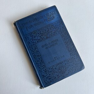 May include: A vintage book titled "Girls and Women" by E. Chester, part of "The Riverside Library For Young People" series. The book has a dark blue cover with ornate black floral designs and text. The book is in good condition.
