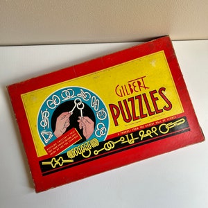 May include: A red and yellow box with the text "GILBERT PUZZLES" and "A PRODUCT FROM THE GILBERT HALL OF SCIENCE". The box features a variety of metal puzzles, including rings, chains, and knots. The text "BAFFLING TWISTERS - THEY TEST YOUR PUZZLE SOLVING SKILL - LOADS OF FUN FOR EVERYONE" is also on the box.