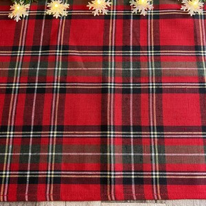 May include: Red and black plaid table runner with a white border. The runner is decorated with a string of white snowflake lights.
