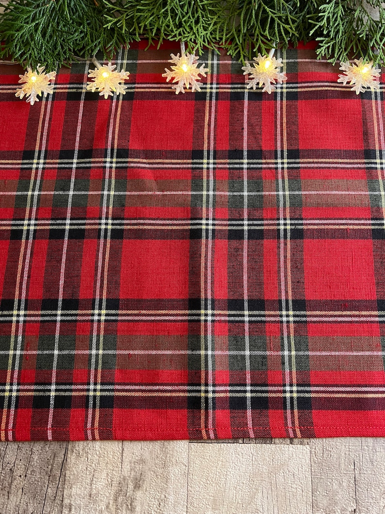 May include: Red and black plaid table runner with a white border. The runner is decorated with a string of white snowflake lights.