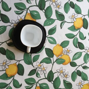 May include: A white ceramic cup with a black saucer, sitting on a white tablecloth with a lemon tree pattern.