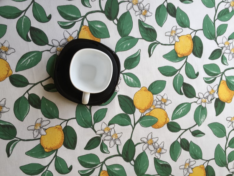 May include: A white ceramic cup with a black saucer, sitting on a white tablecloth with a lemon tree pattern.