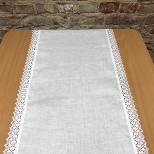 Puede incluir: Un camino de mesa de lino blanco con un delicado borde de encaje blanco en ambos lados. El camino de mesa está colocado sobre una mesa de madera.