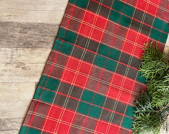 May include: Red and green plaid table runner with a subtle brown plaid pattern. The runner is made of a woven fabric and has a fringed edge.