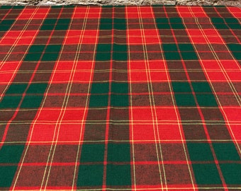 May include: A red, green, and gold plaid fabric. The fabric is a traditional tartan pattern with intersecting lines creating a grid design. The background is a weathered brick wall.