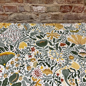 Op de afbeelding: Een close-up van een stof met een botanisch patroon. Het ontwerp heeft een witte achtergrond met een herhalend patroon van bloemen, bladeren en stelen in tinten geel, groen, grijs en bruin. De stof wordt waarschijnlijk gebruikt voor woondecoratie.