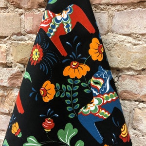 May include: Black kitchen towel with a colorful Swedish Dala horse design. The horses are red, blue, and green with white accents. The background is black with green leaves and orange flowers.