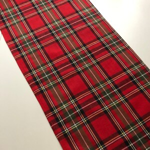 May include: Red and green plaid table runner with a white background. The runner is made of a woven fabric and has a fringed edge.