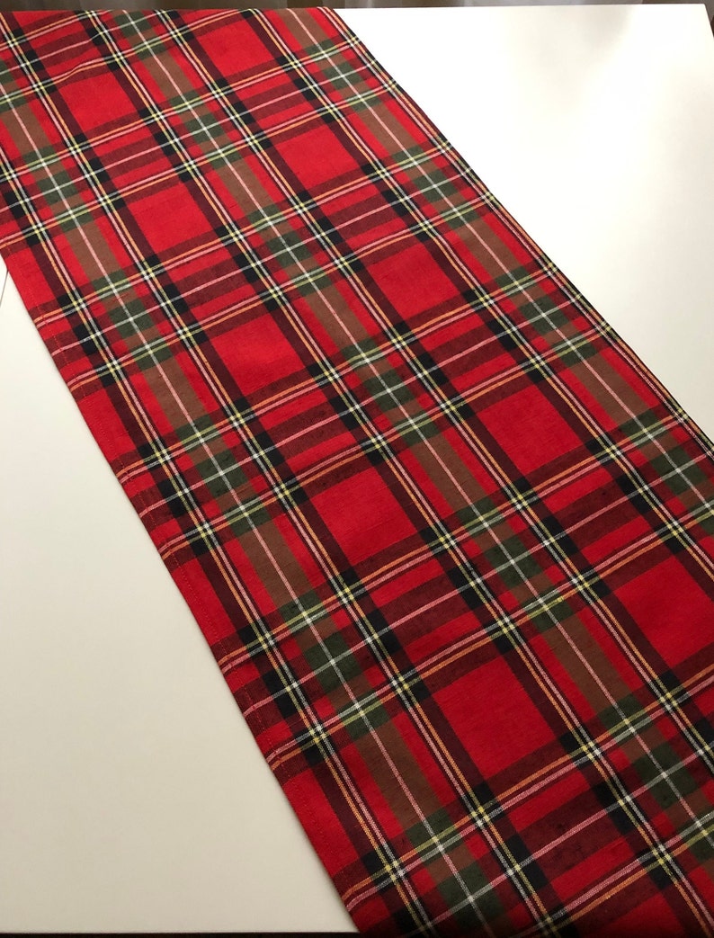 May include: Red and green plaid table runner with a white background. The runner is made of a woven fabric and has a fringed edge.