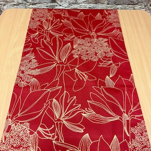 Może przedstawiać: Czerwony bieżnik na stół ze złotym wzorem kwiatowym. Bieżnik przedstawia duże, stylizowane kwiaty i mniejsze, kropkowane elementy kwiatowe. Wzór jest nadrukowany na jednolitym czerwonym tle, tworząc uderzający kontrast. Bieżnik jest prezentowany na jasnym drewnianym stole.