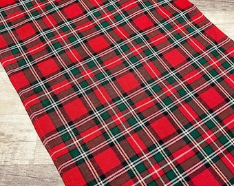 May include: Red plaid table runner with green, white, and brown accents. The fabric has a classic tartan pattern, perfect for holiday or seasonal decor. The runner is made of a woven material.