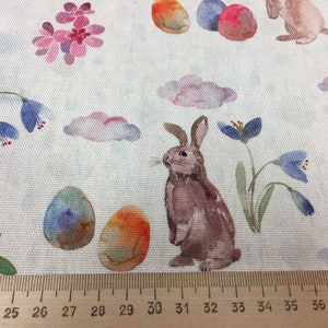 May include: A watercolor fabric design featuring a brown bunny rabbit, blue and purple flowers, and colorful Easter eggs on a white background.