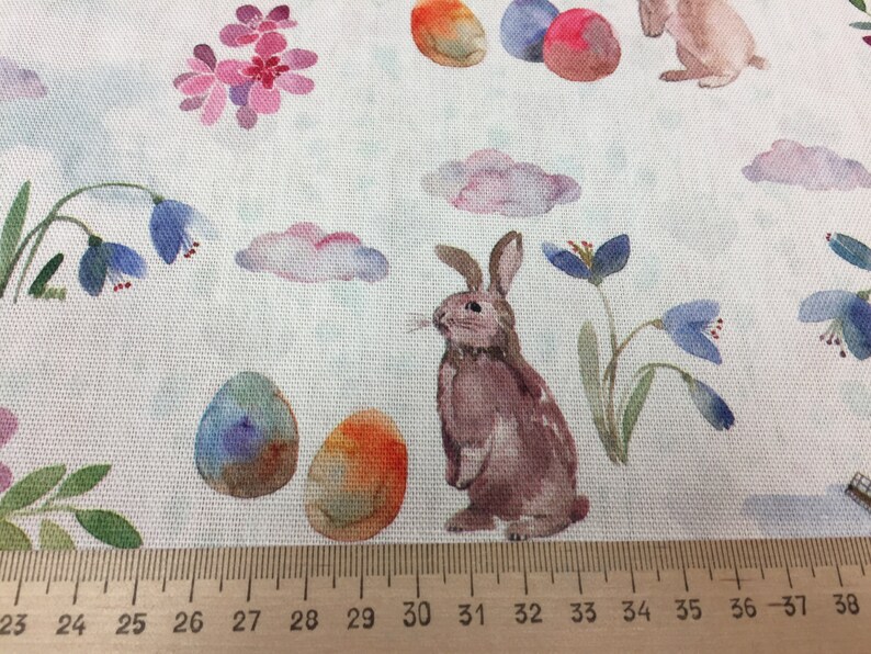 May include: A watercolor fabric design featuring a brown bunny rabbit, blue and purple flowers, and colorful Easter eggs on a white background.