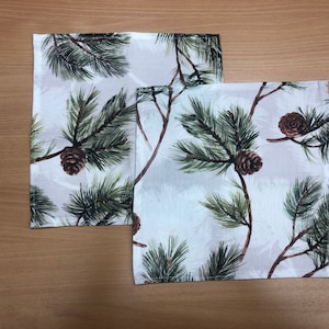 May include: Two white cotton napkins with a pine tree and pine cone design. The napkins have a white background with green pine needles and brown pine cones.