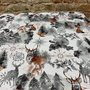 May include: White tablecloth with a winter forest print. The design features deer, foxes, squirrels, trees, and snowflakes in shades of brown, gray, and white.