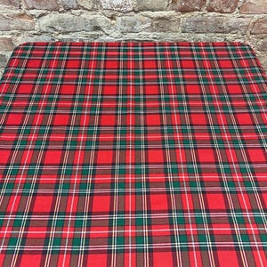 Puede incluir: Un mantel a cuadros rojos con líneas verdes, blancas y marrones. La tela está extendida sobre una mesa, con una pared de ladrillos al fondo. El patrón es un diseño tartán clásico.
