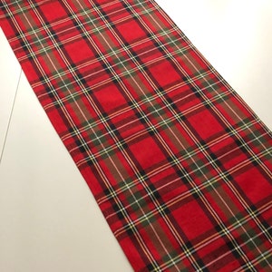 May include: Red and green plaid table runner. The runner has a traditional tartan pattern with a red background and green, black, and yellow stripes.