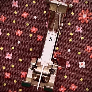 Puede incluir: Un prensatelas plateado para máquina de coser con el número 5 en una etiqueta blanca. El prensatelas tiene una guía para la aguja y está sobre una tela marrón con estampado floral. Diseñado para coser.
