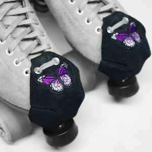 May include: A pair of grey roller skates with black toe stops. Each toe stop has a purple butterfly embroidered on a black fabric cover.