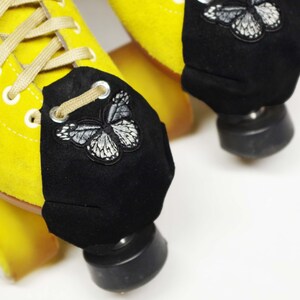 May include: Close-up of yellow roller skates with black toe caps. Each cap features a white and black embroidered butterfly. The skates have tan laces and black wheels, set against a white background.