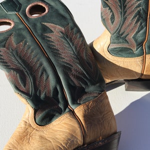May include: A pair of tan leather cowboy boots with green leather tops and intricate stitching. The boots have a pointed toe and a low heel.