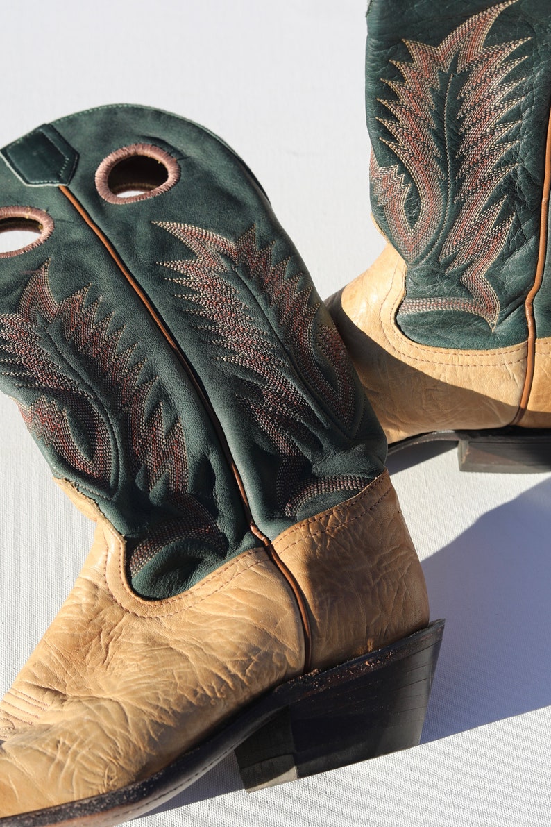 May include: A pair of tan leather cowboy boots with green leather tops and intricate stitching. The boots have a pointed toe and a low heel.