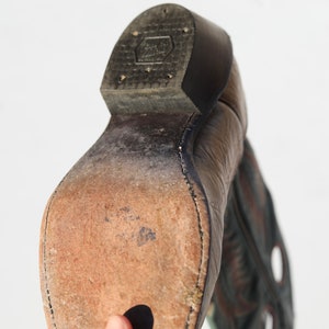 May include: Close-up of the bottom of a brown leather cowboy boot with a black leather heel. The boot has a stitched seam around the sole and a worn leather insole.