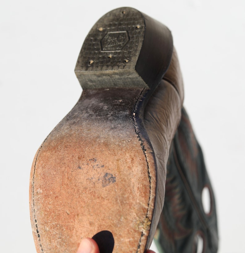 May include: Close-up of the bottom of a brown leather cowboy boot with a black leather heel. The boot has a stitched seam around the sole and a worn leather insole.