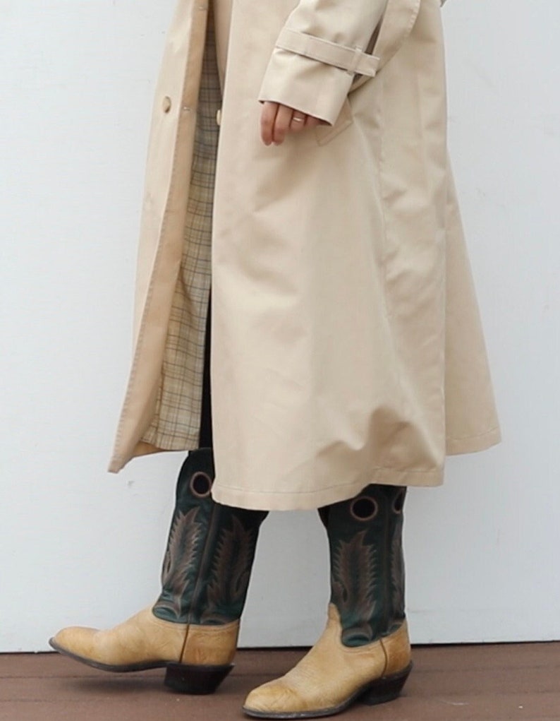 May include: A beige trench coat with a plaid lining and a pair of tan cowboy boots with green and black embroidery.