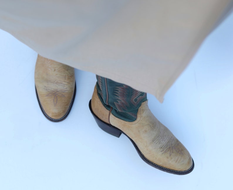 May include: A pair of tan cowboy boots with green embroidered details. The boots have a pointed toe and a low heel.