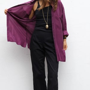 May include: A long-sleeved purple button-down shirt worn over a black tank top and black trousers. The outfit includes a long gold-toned necklace with an oval pendant and black leather boots. The shirt features a collar and a chest pocket.